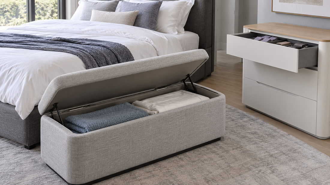 Open gray upholstered storage bench holding bedding beside a white dresser with an open drawer in a contemporary bedroom.