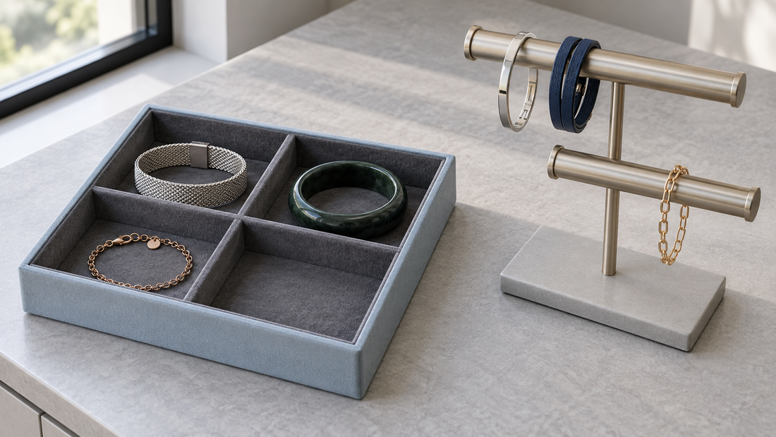 Bracelets arranged in a four-compartment blue-gray tray beside a two-rod metal bracelet stand on a gray dressing table.