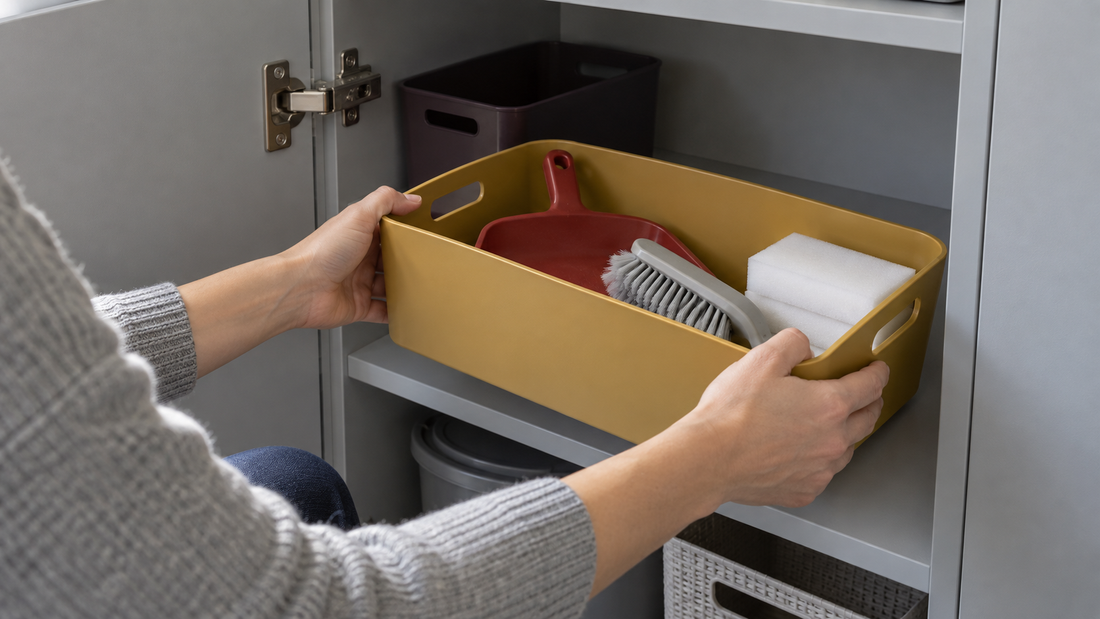 Hands removing an ochre storage bin containing cleaning tools from a gray cabinet beside an open door and visible hinge.