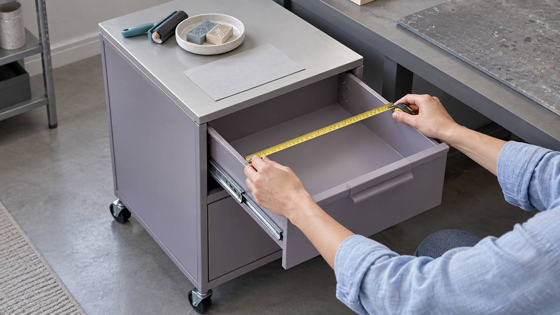 A person measures the interior width of a drawer trolley with craft supplies on its mostly clear upper surface.