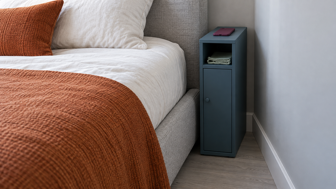 A slim blue nightstand fits between a bed and the wall, holding a phone on top and a folded cloth on its open shelf.