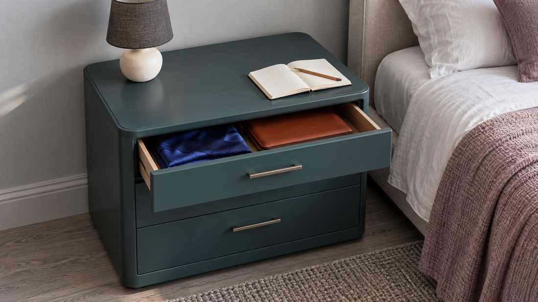 A teal bedside chest offers clear tabletop space beside a lamp and open notebook, with a scarf and leather folio stored in its open drawer.