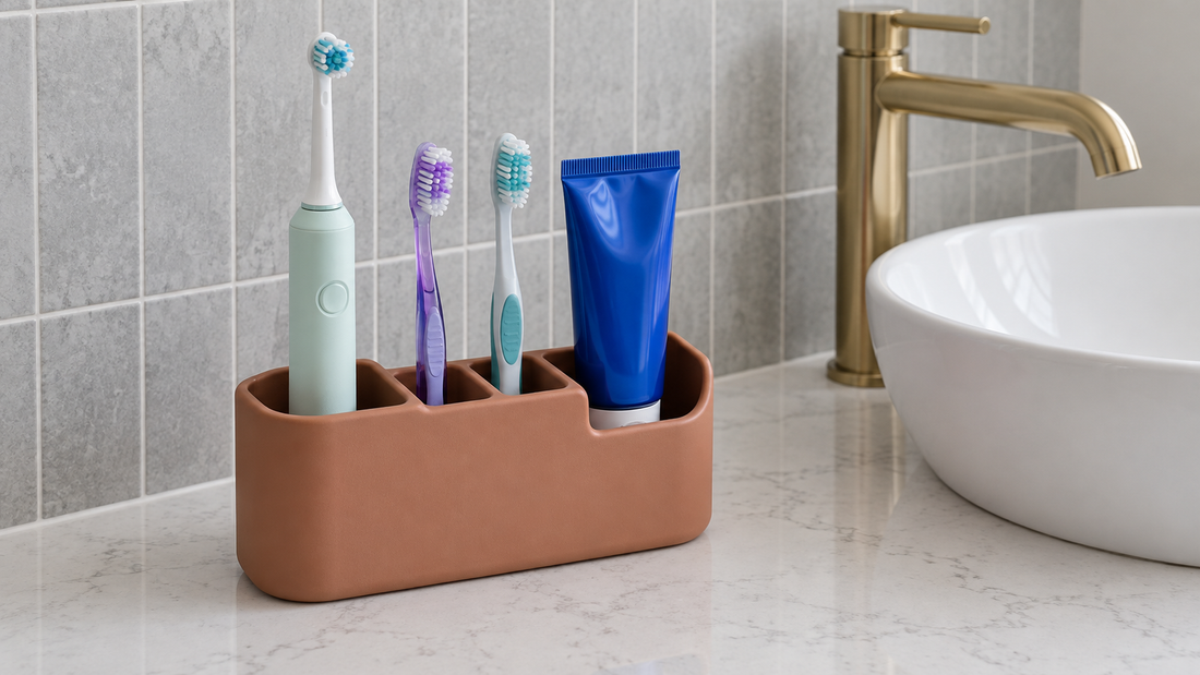 A terracotta organizer holds an electric toothbrush, two manual toothbrushes, and a blue toothpaste tube in differently sized sections beside a bathroom sink.