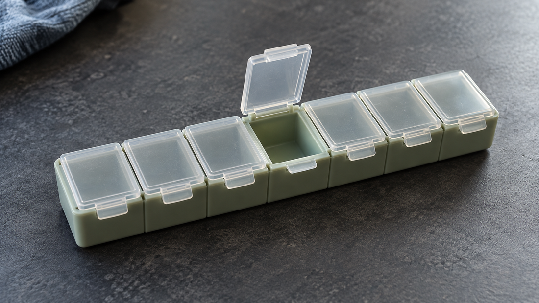 A sage-green weekly pill organizer has its center lid open while all six neighboring compartments remain closed.